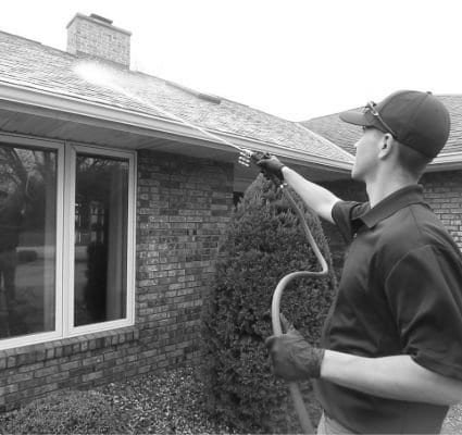 A worker from Coronado Window Cleaning sprays a roof, cleaning it with a pressure washer. The house is brick with large windows, and a green bush is visible in the foreground. San Diego Window Cleaning services.