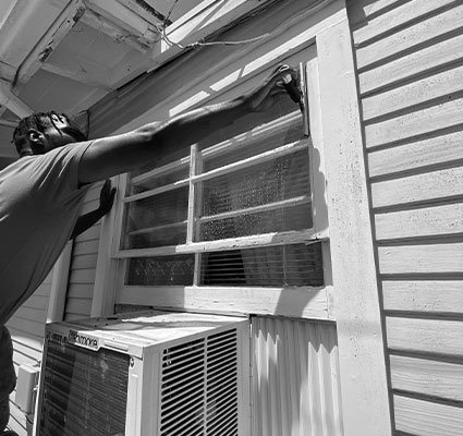 A man performing Coronado Window Cleaning on an exterior window with an AC unit below. He's using a brush to clean the windowpane. San Diego Window Cleaning services at work.