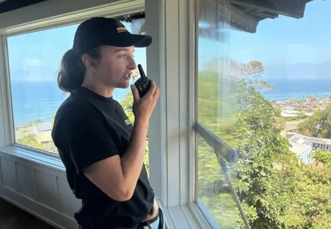 A window cleaner communicates via walkie-talkie while cleaning windows on a multi-story Point Loma home. Ocean views and lush greenery are visible through the sparkling glass.