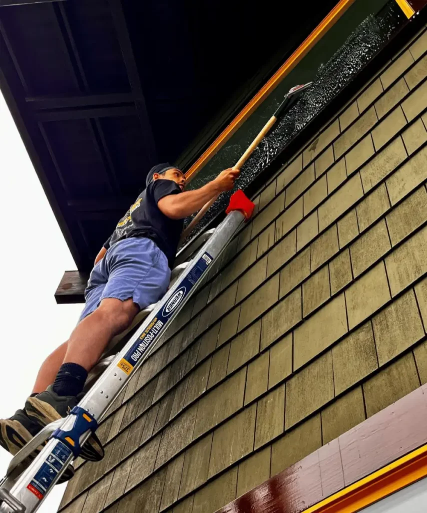 A man on a ladder performs advanced window cleaning on a multi-story home in Point Loma. He uses a squeegee to clean a high window, showcasing the meticulous work required for high-end home maintenance.