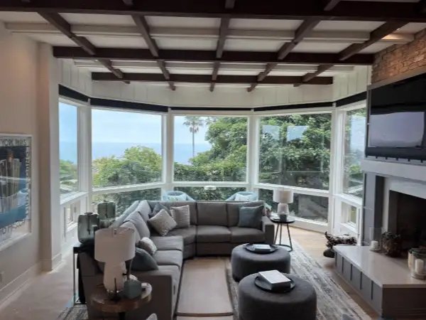 Bright, airy living room with a large bay window showcasing an ocean view. A gray sectional sofa and ottomans sit atop a patterned rug. This Point Loma home features advanced window cleaning for optimal views.