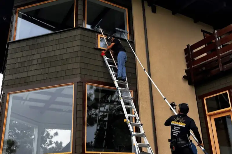 Two window cleaners perform advanced window cleaning on a multi-story home in Point Loma. One uses a ladder, the other a long pole with a squeegee. Sunshine Cleaning logo visible.