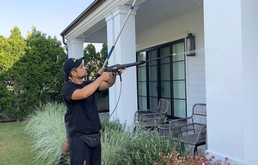 Man power washing a white house exterior with a Ryobi pressure washer. He wears a black t-shirt and cap, demonstrating how to wash your house the right way.