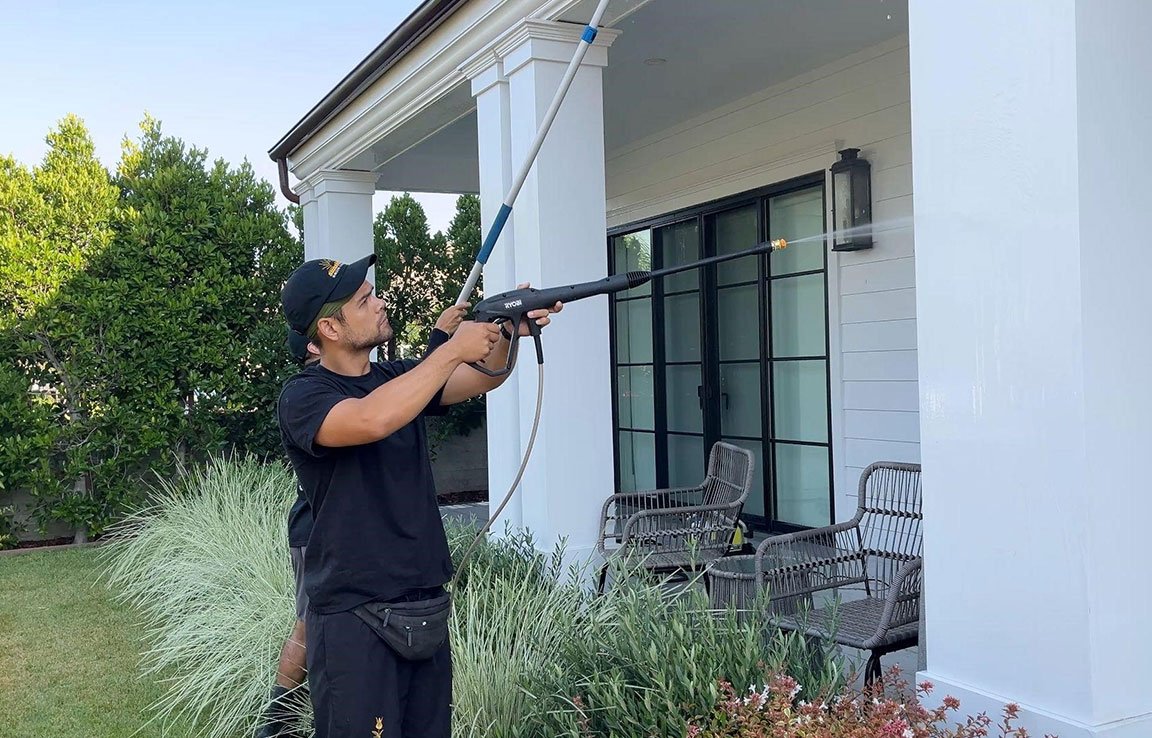 Man power washing a white house exterior with a Ryobi pressure washer. He wears a black t-shirt and cap, demonstrating how to wash your house the right way.