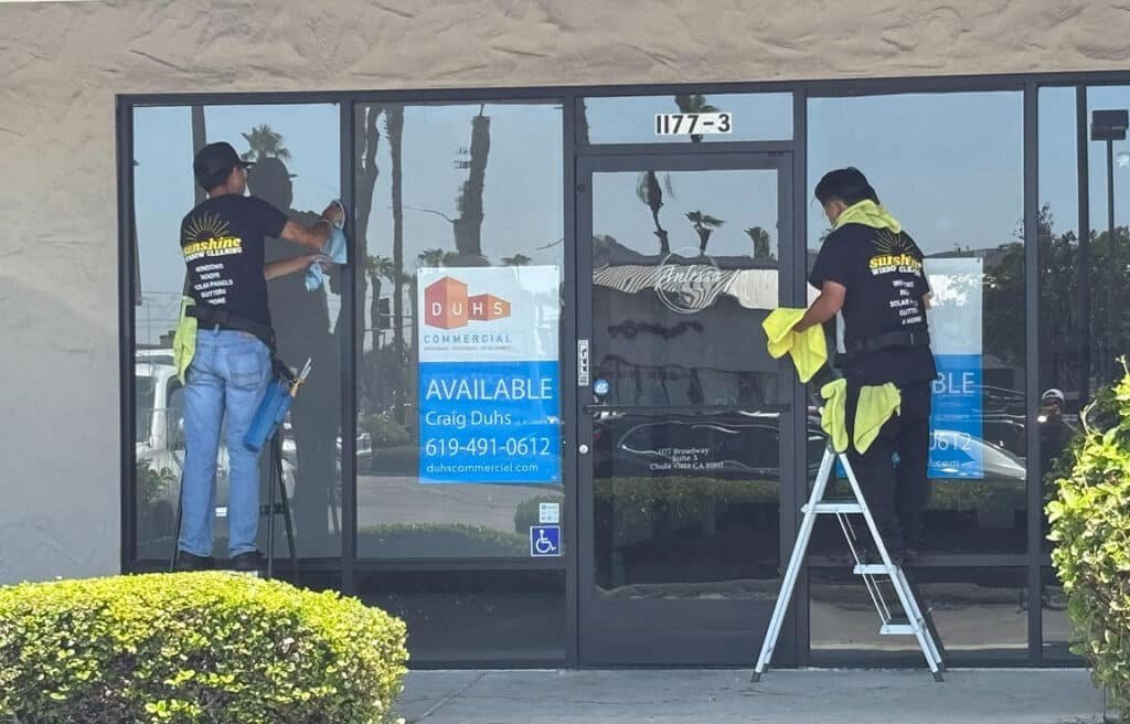 Two window cleaners from Sunshine Window Cleaning are cleaning the storefront windows of a commercial property in San Diego. The windows have a commercial real estate "Available" sign.