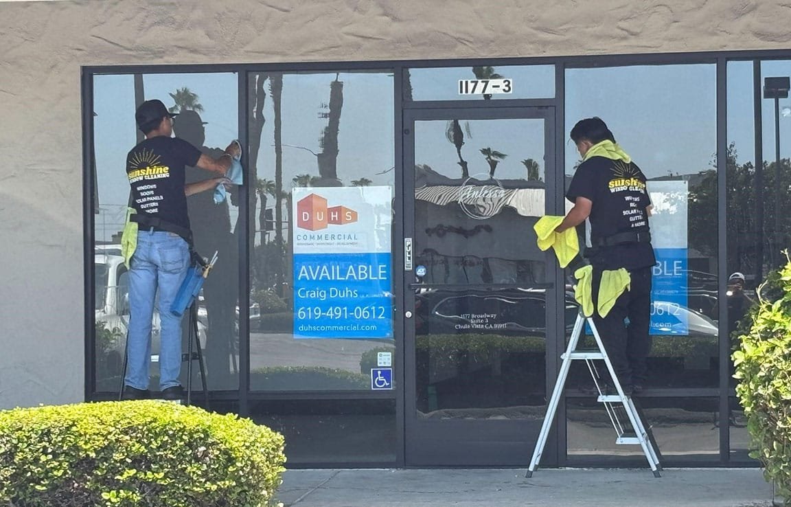 Two window cleaners from Sunshine Window Cleaning are cleaning the storefront windows of a commercial property in San Diego. The windows have a commercial real estate "Available" sign.